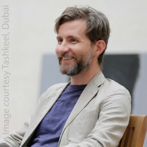 Image of Gallery Director, Flounder Lee. He is a white cis male with greyish beard, medium length brown hair, he is smiling looking to the side and wearing a tan blazer and a blue tee shirt.